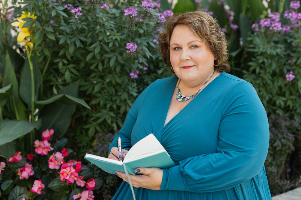 Cindy Tschosik in front of flowering bushes, holding a notebook and a pen